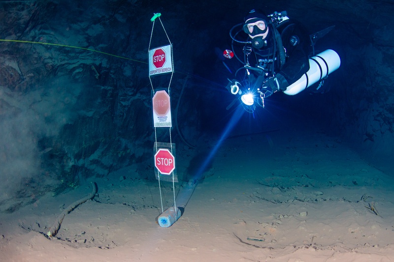 Sidemount cave diver hovering beside underwater stop signs marking a restricted zone inside a flooded mine tunnel at Kleinenbremen.